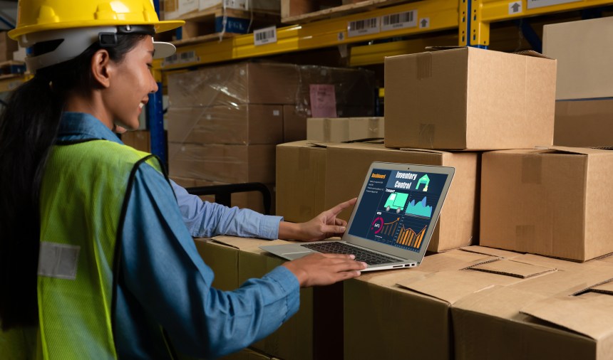 Woman in safety gear analyzes inventory data on a laptop in a warehouse with stacked boxes.