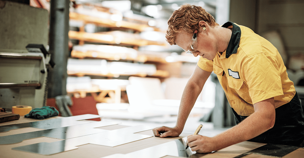 Man in a yellow shirt measuring and marking a wooden workbench in a workshop