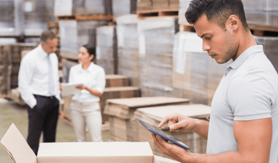 Warehouse worker using a tablet, with colleagues discussing in the background among boxes.