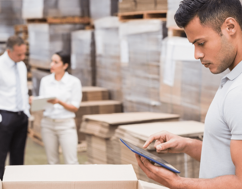 Warehouse worker using a tablet, with colleagues discussing in the background among boxes.
