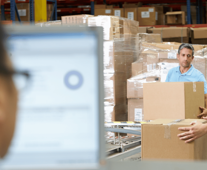 Two workers handle boxes in a warehouse, with a computer monitor visible in the foreground.