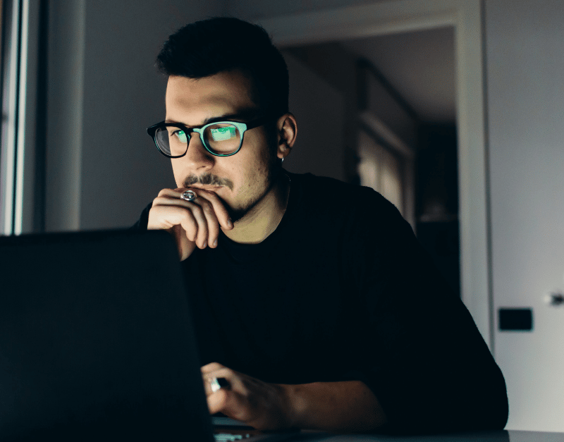 Man in glasses looking thoughtfully at a laptop screen in dim lighting