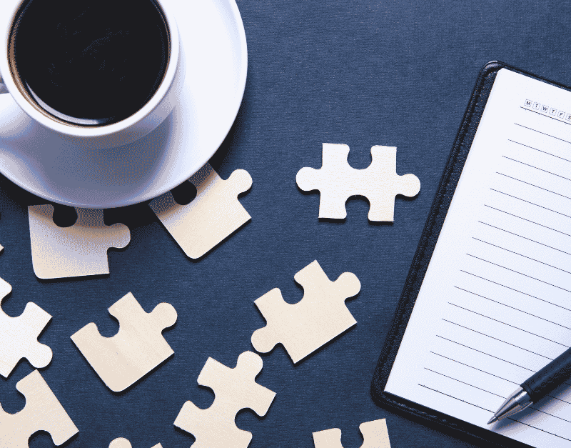 A coffee cup sits beside scattered puzzle pieces and an open notebook with a pen, set against a dark background.