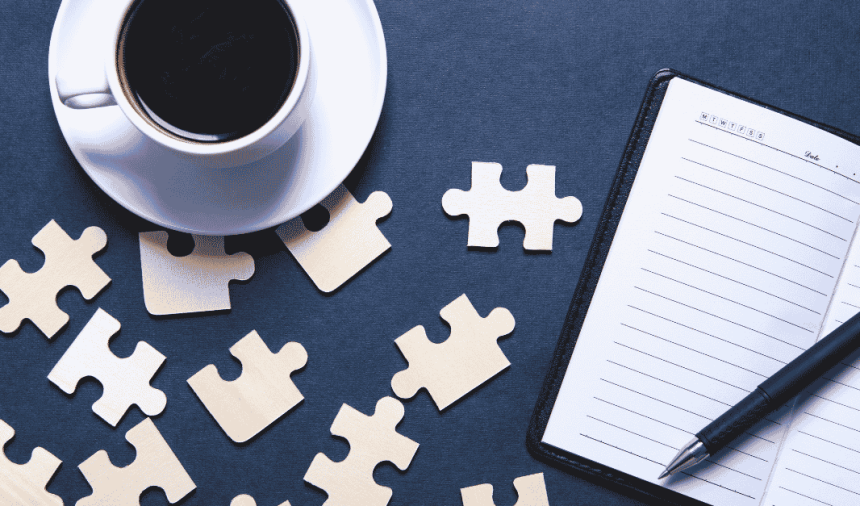 A coffee cup sits beside scattered puzzle pieces and an open notebook with a pen, set against a dark background.