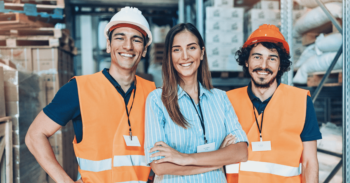 Three smiling workers in safety vests pose together in a warehouse filled with stacked boxes.