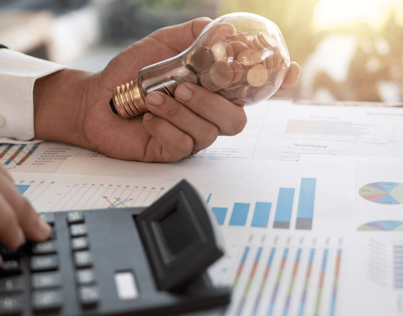 A hand holds a lightbulb filled with coins above financial reports and a calculator.