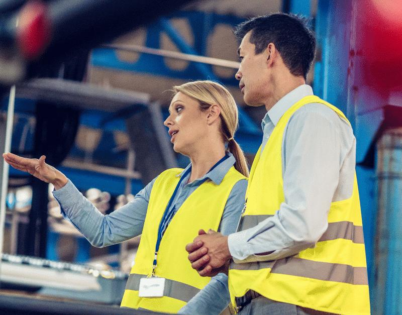 Two individuals in safety vests engage in conversation in a warehouse setting.