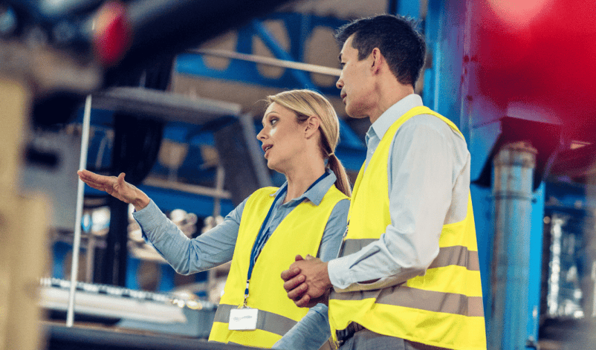 Two individuals in safety vests engage in conversation in a warehouse setting.