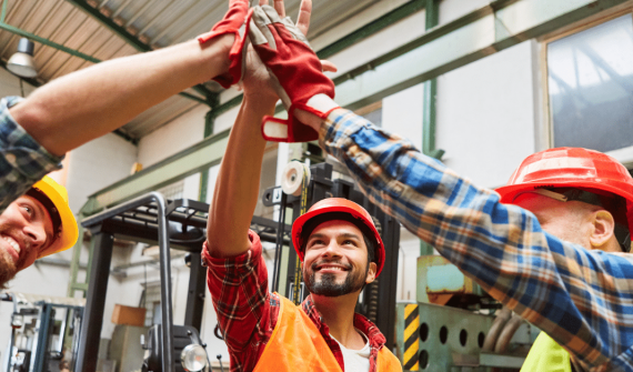 Three construction workers in hard hats celebrate with a high-five in an industrial setting.