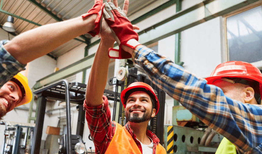 Three construction workers in hard hats celebrate with a high-five in an industrial setting.