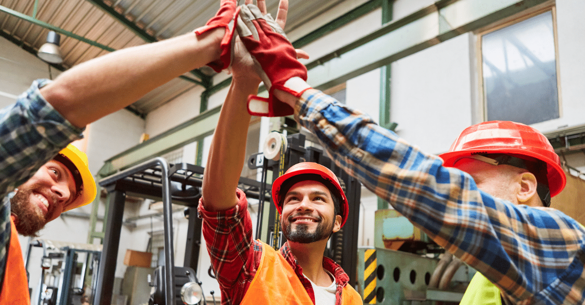Three construction workers in hard hats celebrate with a high-five in an industrial setting.