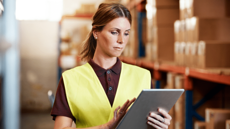 Woman in a yellow safety vest uses a tablet in a warehouse filled with stacked boxes.