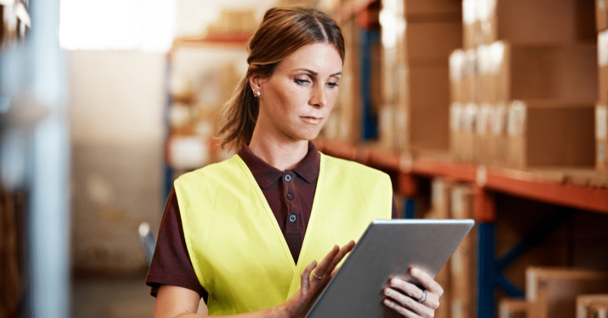 Woman in a yellow safety vest uses a tablet in a warehouse filled with stacked boxes.