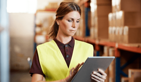 Woman in a yellow safety vest uses a tablet in a warehouse filled with stacked boxes.