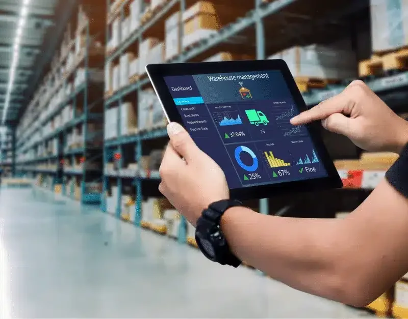 Person uses a tablet displaying warehouse management data in a storage facility aisle.
