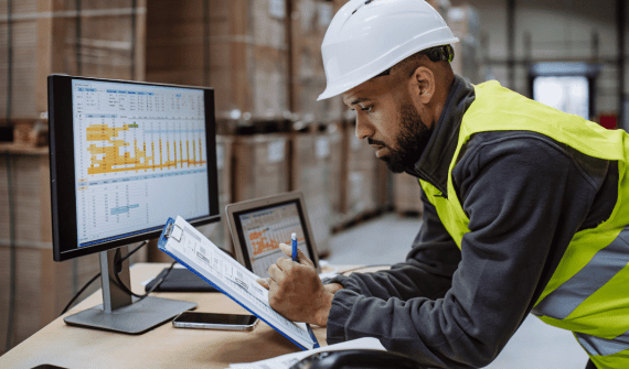 A man in a hard hat and high-visibility vest reviews data on a clipboard and computer.