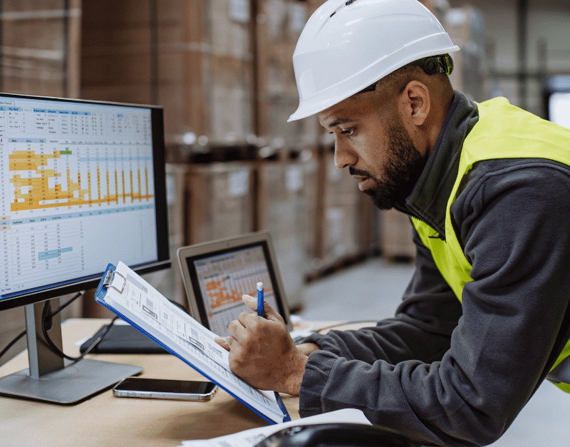 A man in a hard hat and high-visibility vest reviews data on a clipboard and computer.