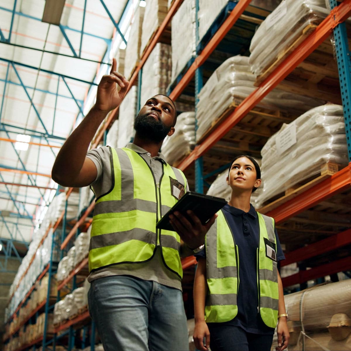 Warehouse workers in safety vests looking up at tall racking filled with wrapped pallets