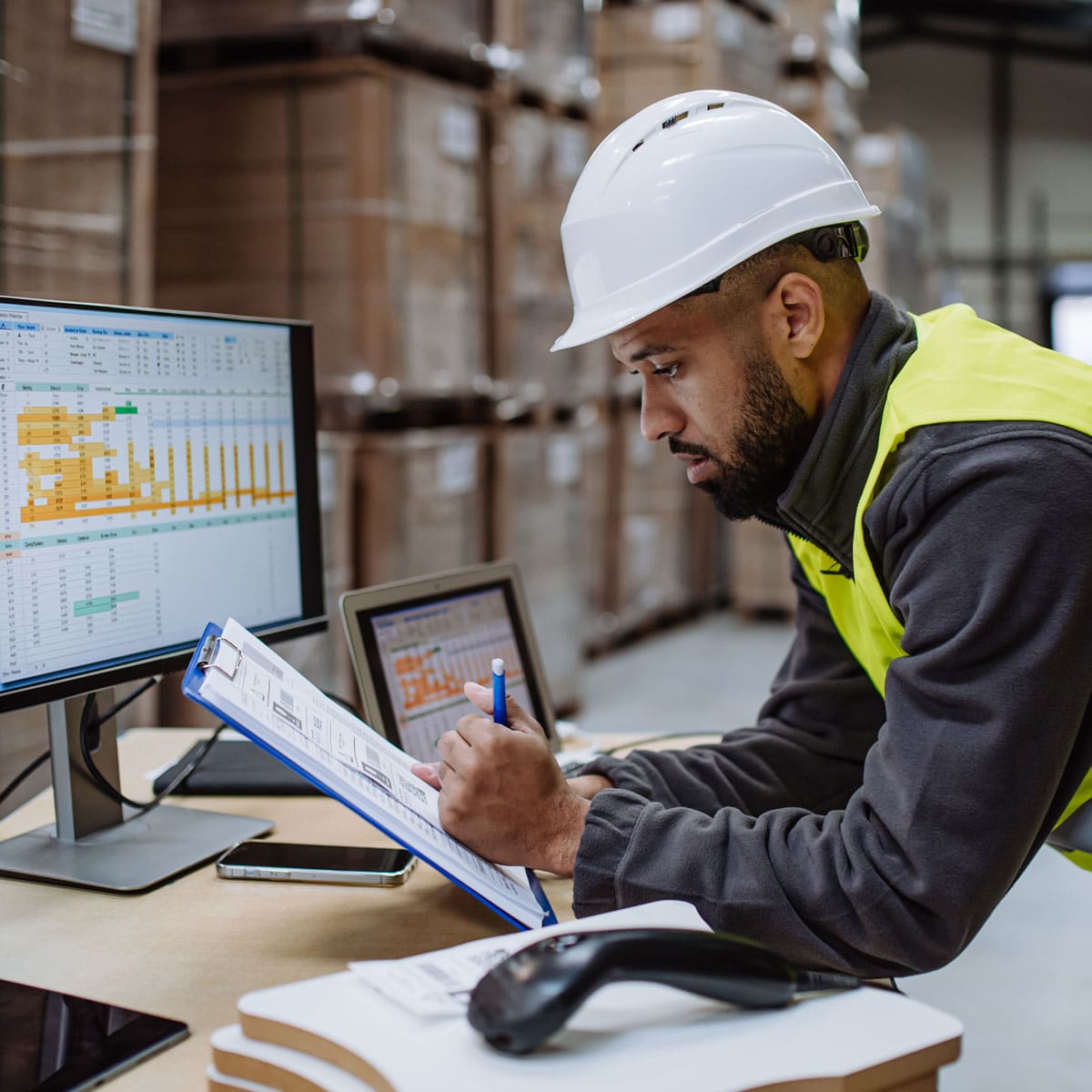 Worker in a hard hat filling out a checklist on a clipboard near computer monitors