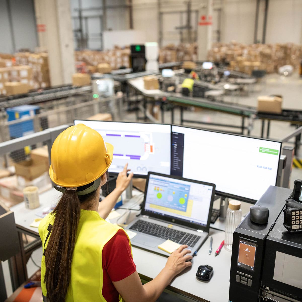 Worker in a hard hat reviewing data across multiple monitors in a warehouse
