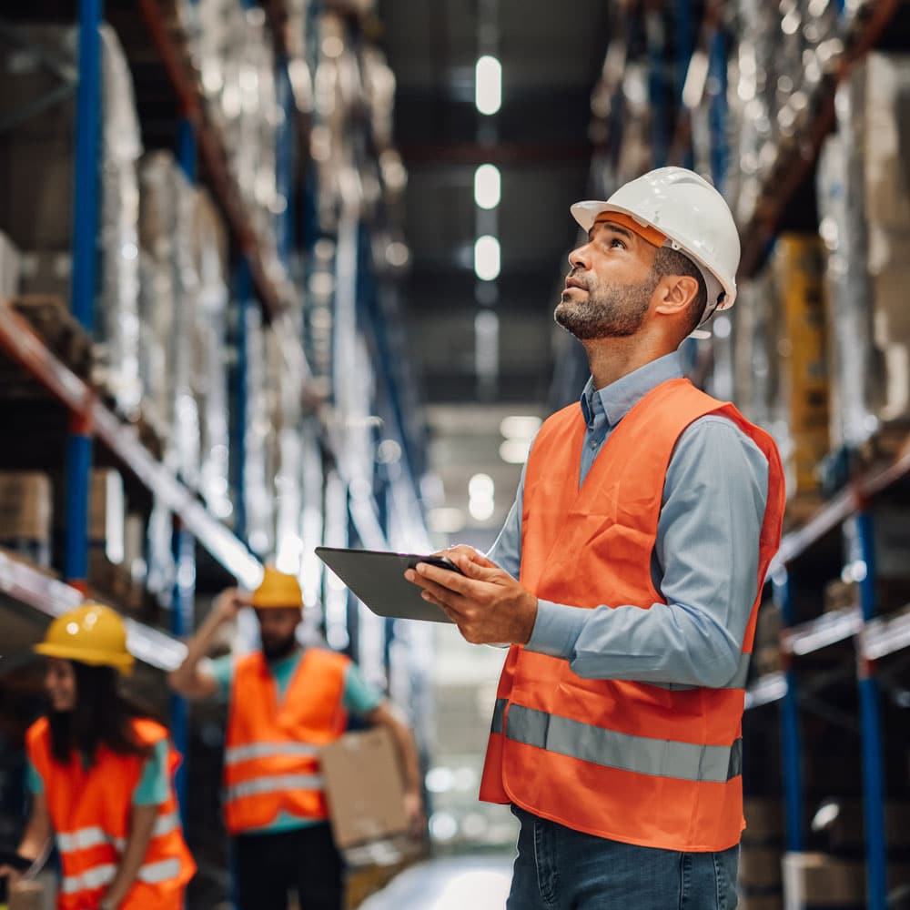 Warehouse worker in a hard hat holding a tablet, looking up at tall storage shelving