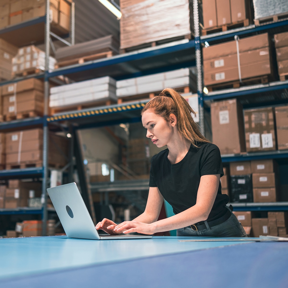 Woman working on a laptop in a warehouse surrounded by boxes on shelves