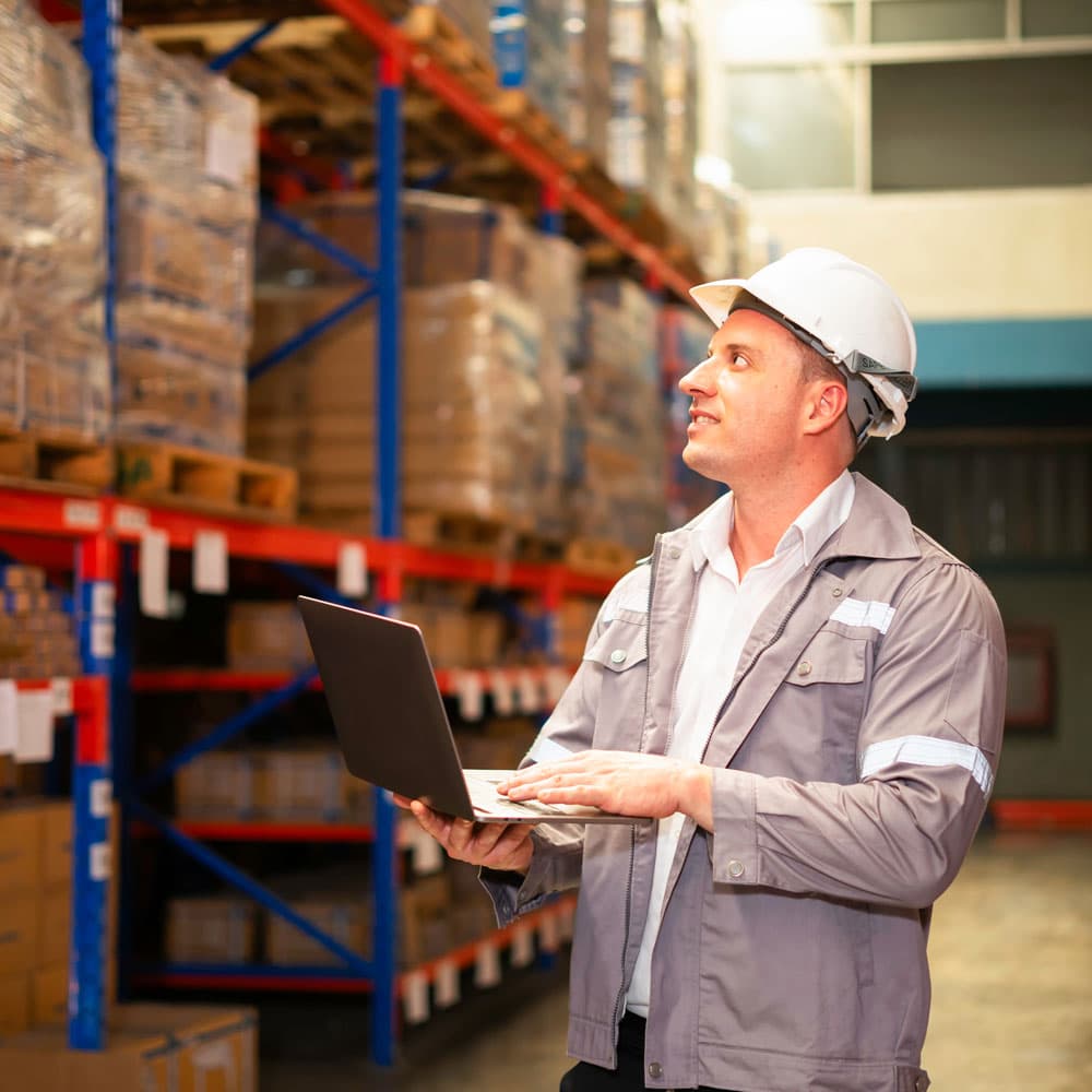 Warehouse worker in a hard hat looking up while holding a laptop